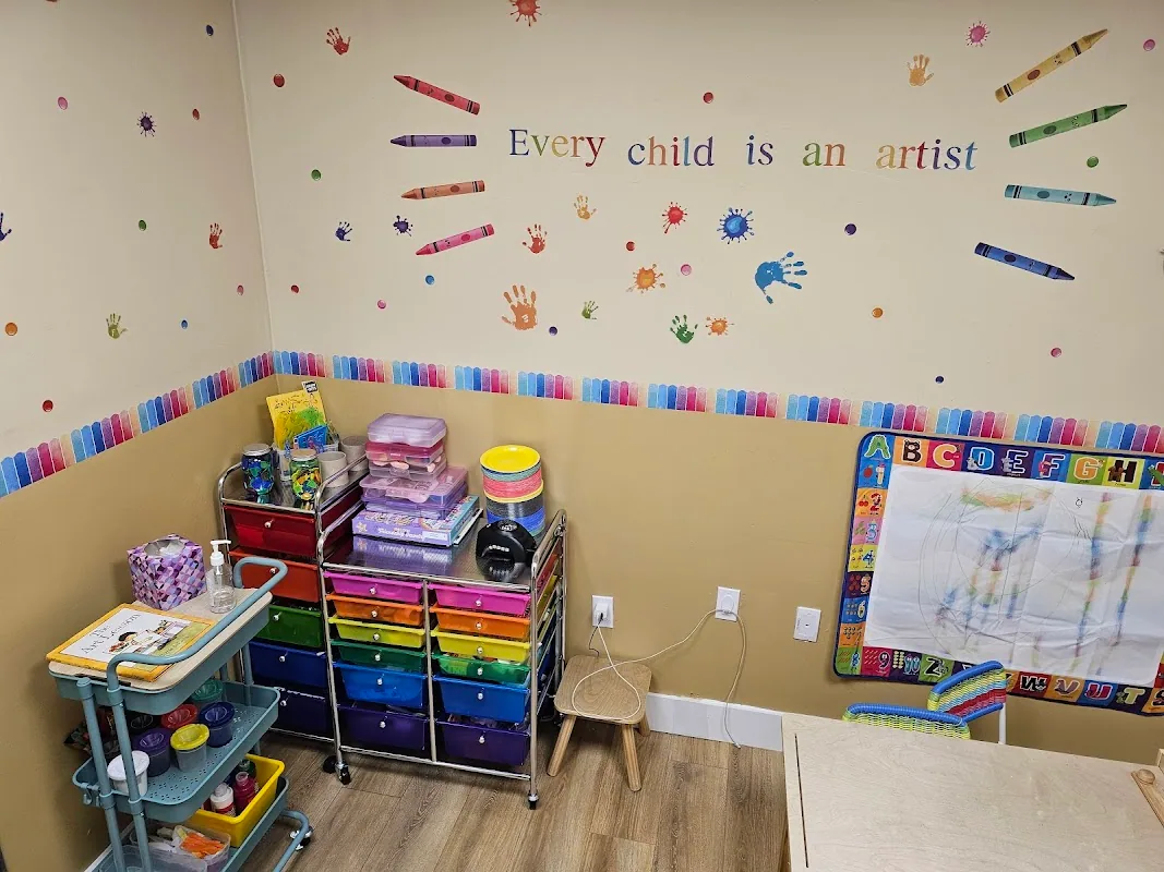 Art room with a colorful 'Every child is an artist' mural, rainbow crayon wall decals, an art-supplies cart with rainbow-colored drawers, and a children's alphabet whiteboard