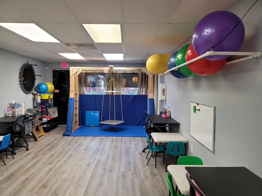 Pediatric sensory gym with therapy balls of different colors mounted along the ceiling, a mini trampoline, a platform swing over a blue mat, and small tables and chairs for children