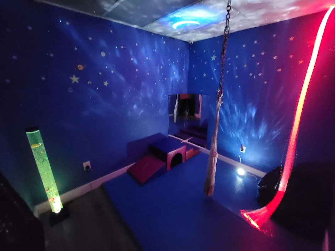 Sensory integration room with deep blue walls and ceiling decorated with stars and planets, a bubble tube lamp, soft foam play shapes on a blue mat, and a hanging swing under a starlight projector