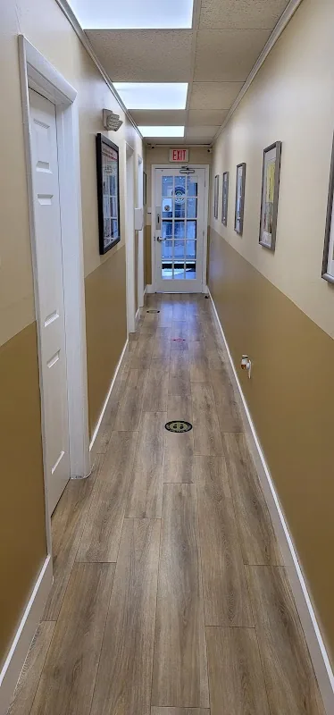 Interior hallway of the clinic with wood-look flooring, framed wall art on each side, an illuminated EXIT sign, and a glass entry door at the end